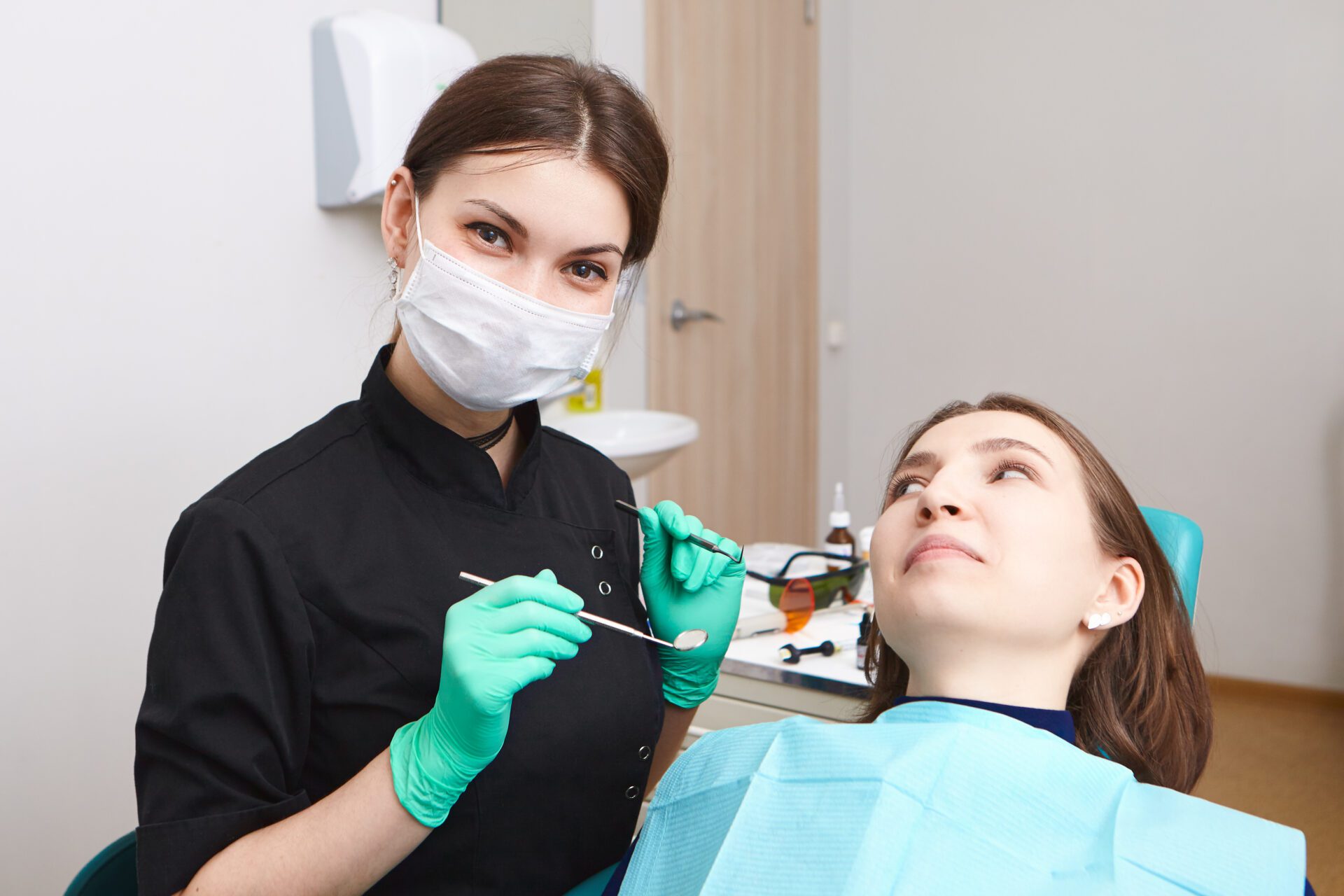 Indoor shot of skillful confident young female dentist wearing exam gloves and white mask holding metal probe and dental miror, ready to examine oral cavity of woman patient sitting in chair Indoor shot of skillful confident young female dentist wearing exam gloves and white mask holding metal probe and dental miror, ready to examine oral cavity of woman patient sitting in chair